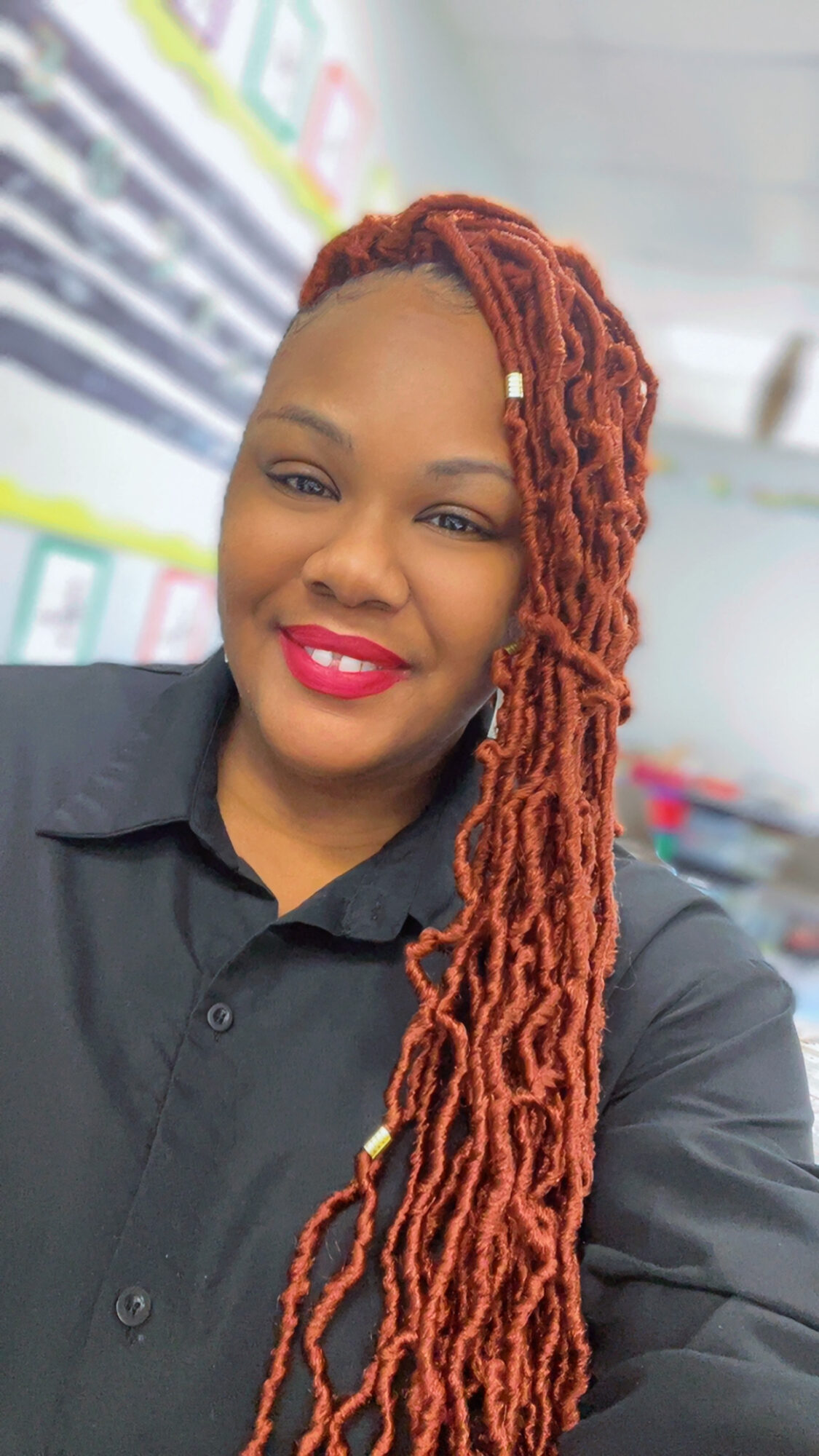 Woman with long, red dreadlocks, smiling, wearing a black shirt, in a store with shelves in background.