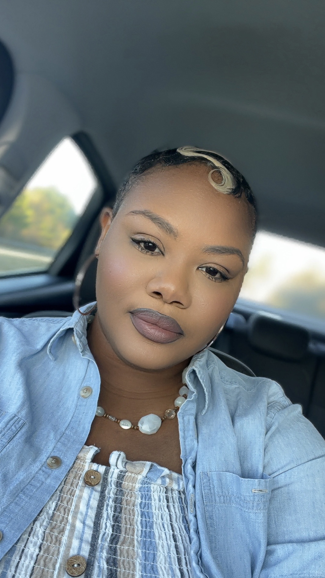 Woman with short hair and makeup, wearing a denim shirt and necklace, sitting in a car.