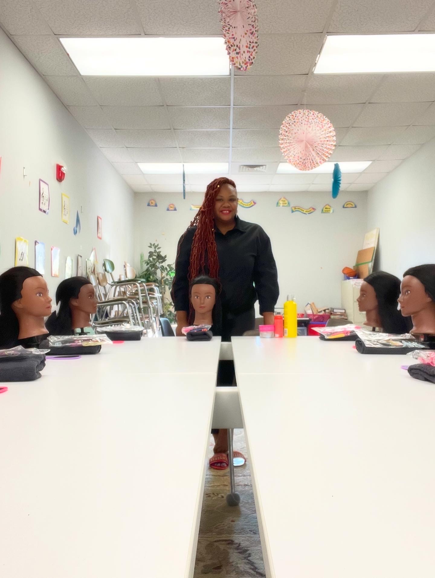 Woman standing at head of classroom with mannequin heads on tables, colorful decorations hanging from ceiling, and classroom supplies visible.