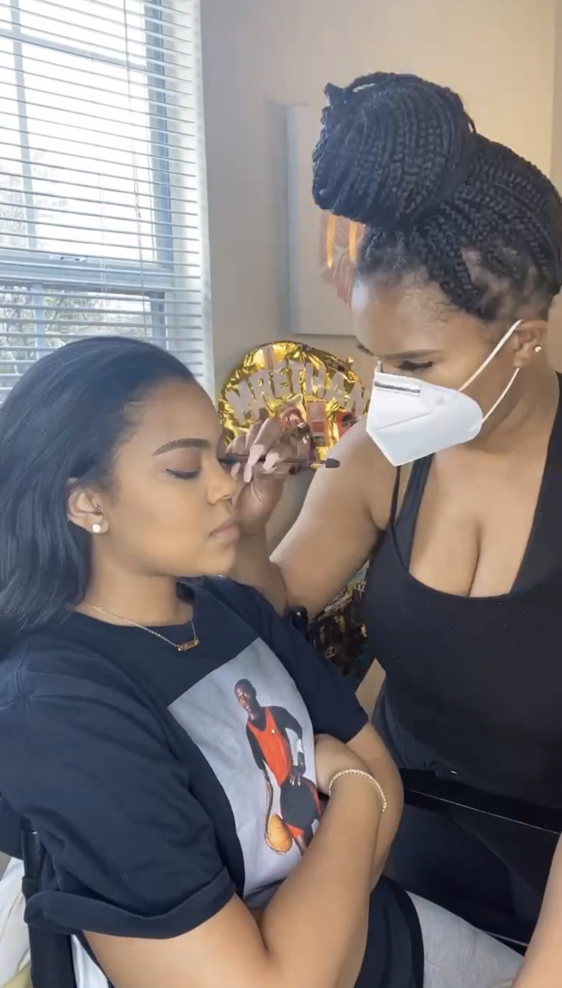 A woman with braided hair and a face mask applies makeup to a young woman with long dark hair, sitting with eyes closed.
