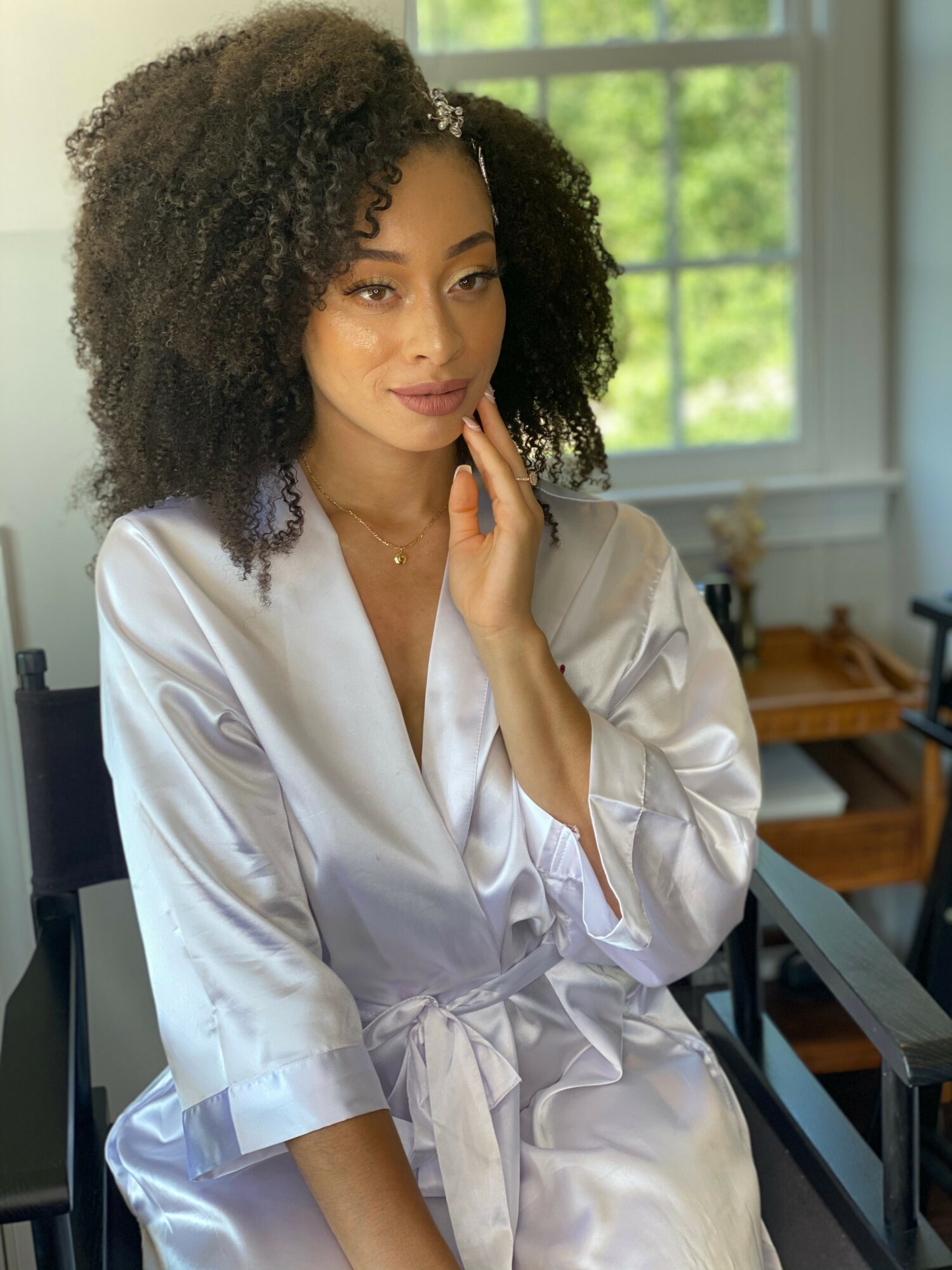 Woman with curly hair in a white satin robe sitting indoors near a window with green trees outside
