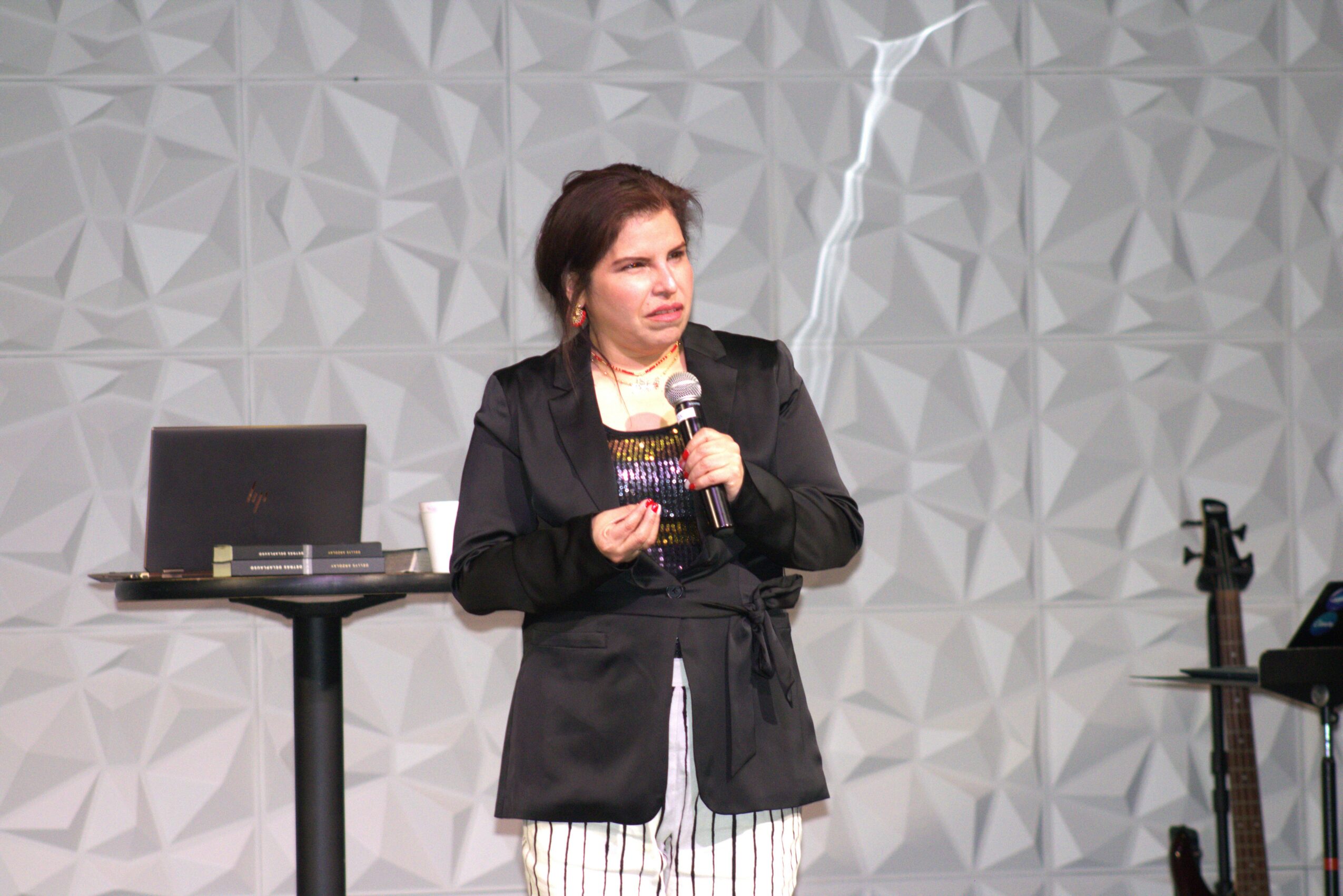 Woman speaking into a microphone on stage with a laptop and a guitar nearby, against a geometric patterned background.