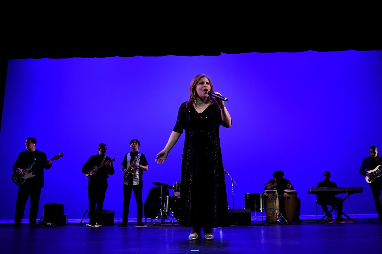 A woman sings into a microphone on stage with a blue background, accompanied by five musicians playing instruments.