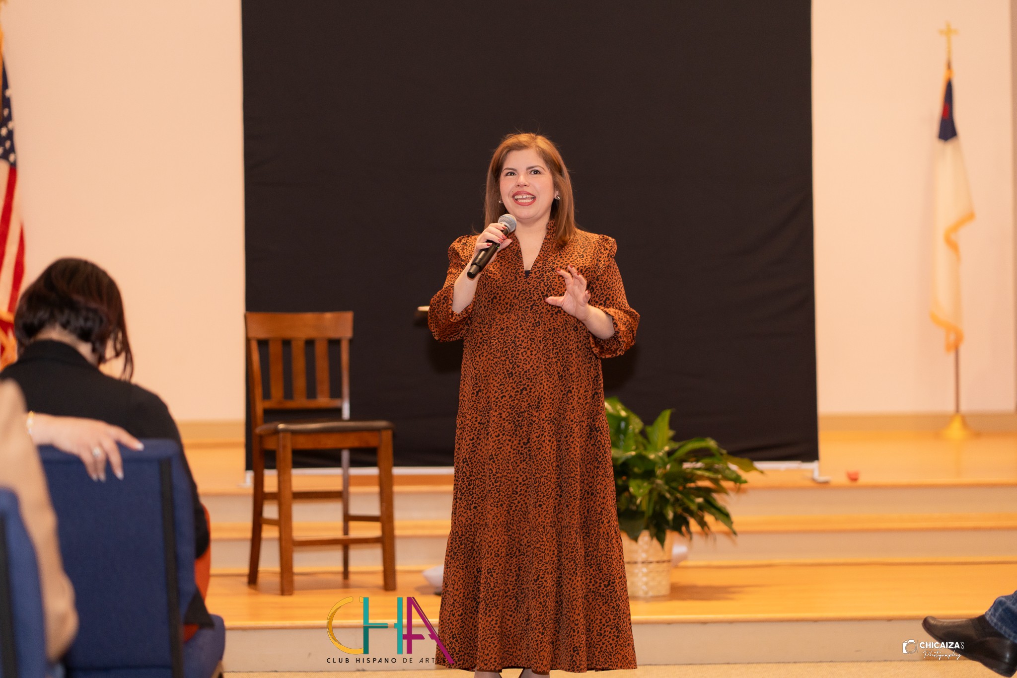 Woman speaking into microphone on stage with chair and plant, flags in background, audience in foreground