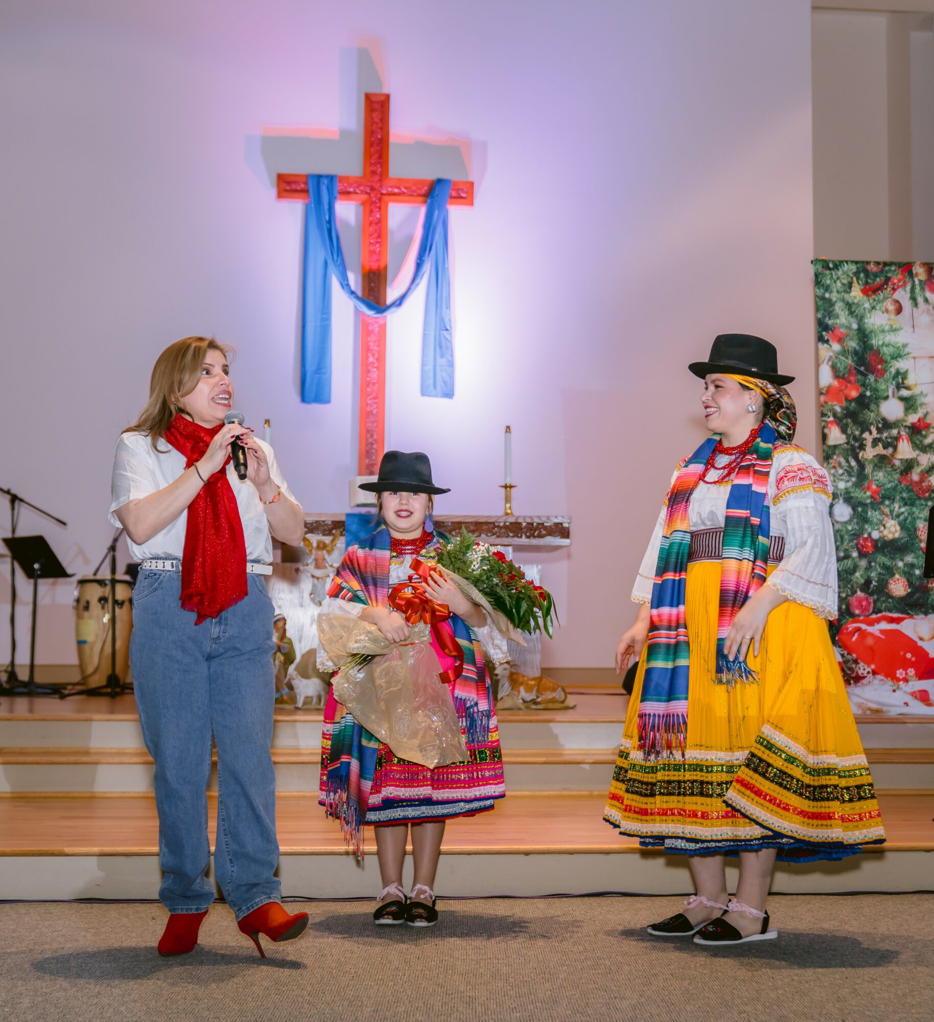 Three women in colorful traditional dresses and scarves stand on a stage with a cross and floral backdrop, engaging in conversation.
