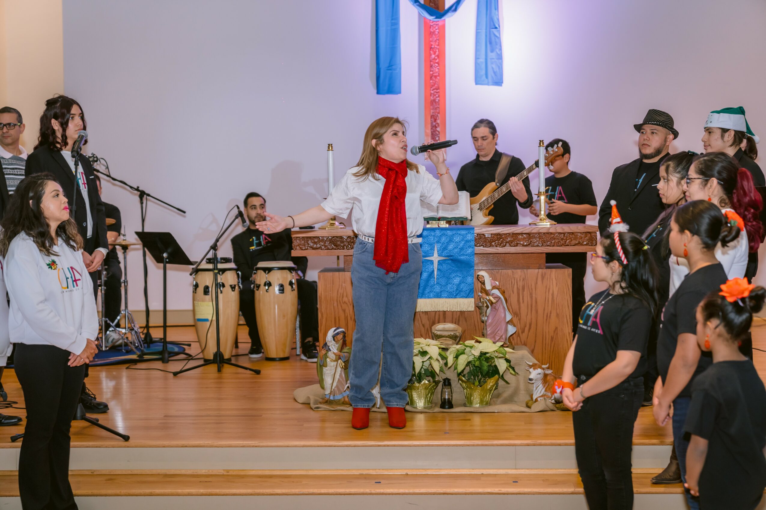 Young girl with red scarf and blue jeans stands on stage with musical instruments and a group of people, some singing and playing instruments.