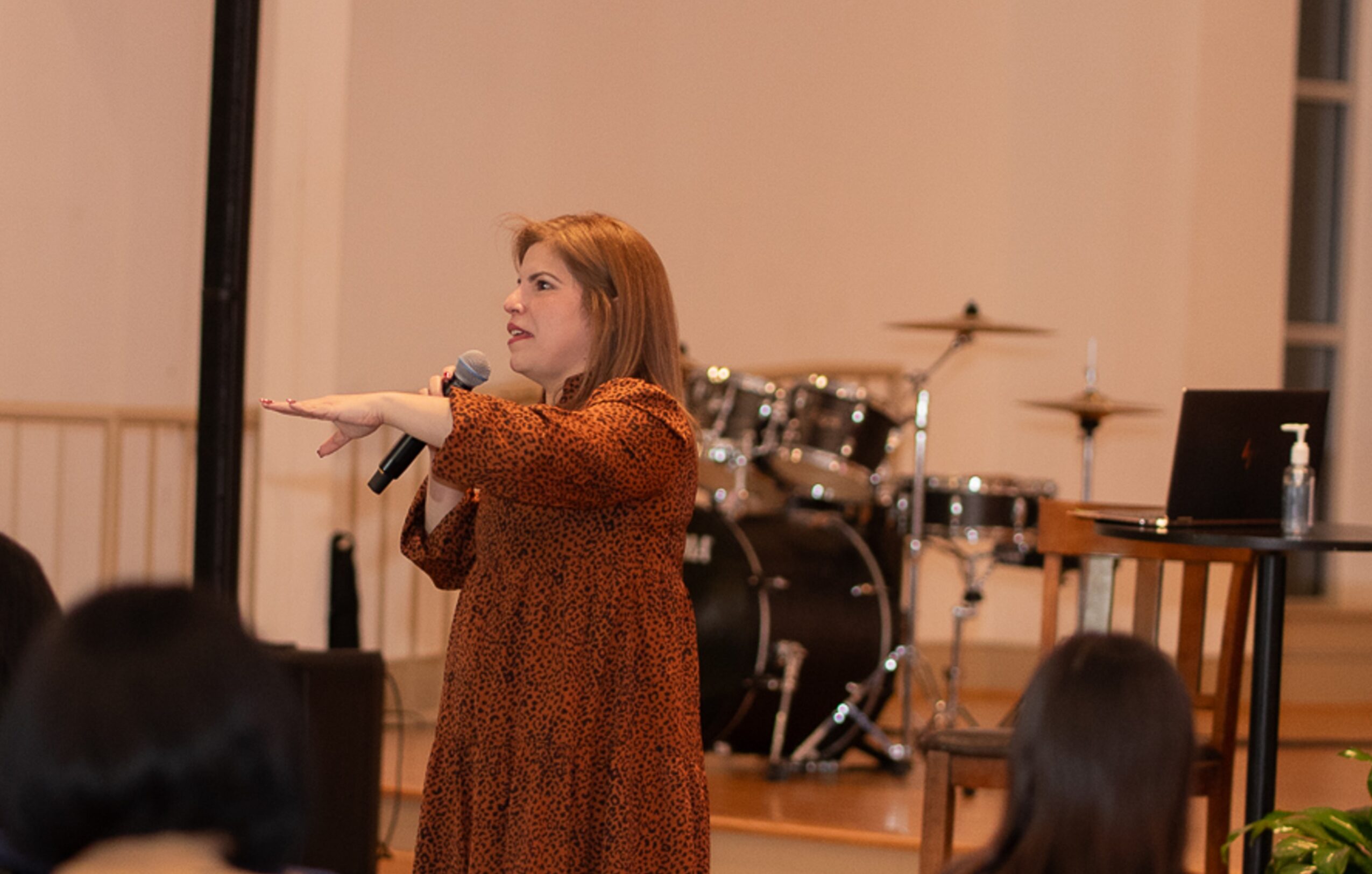 A woman with red hair speaks into a microphone, gesturing with her right hand, in a room with musical instruments.
