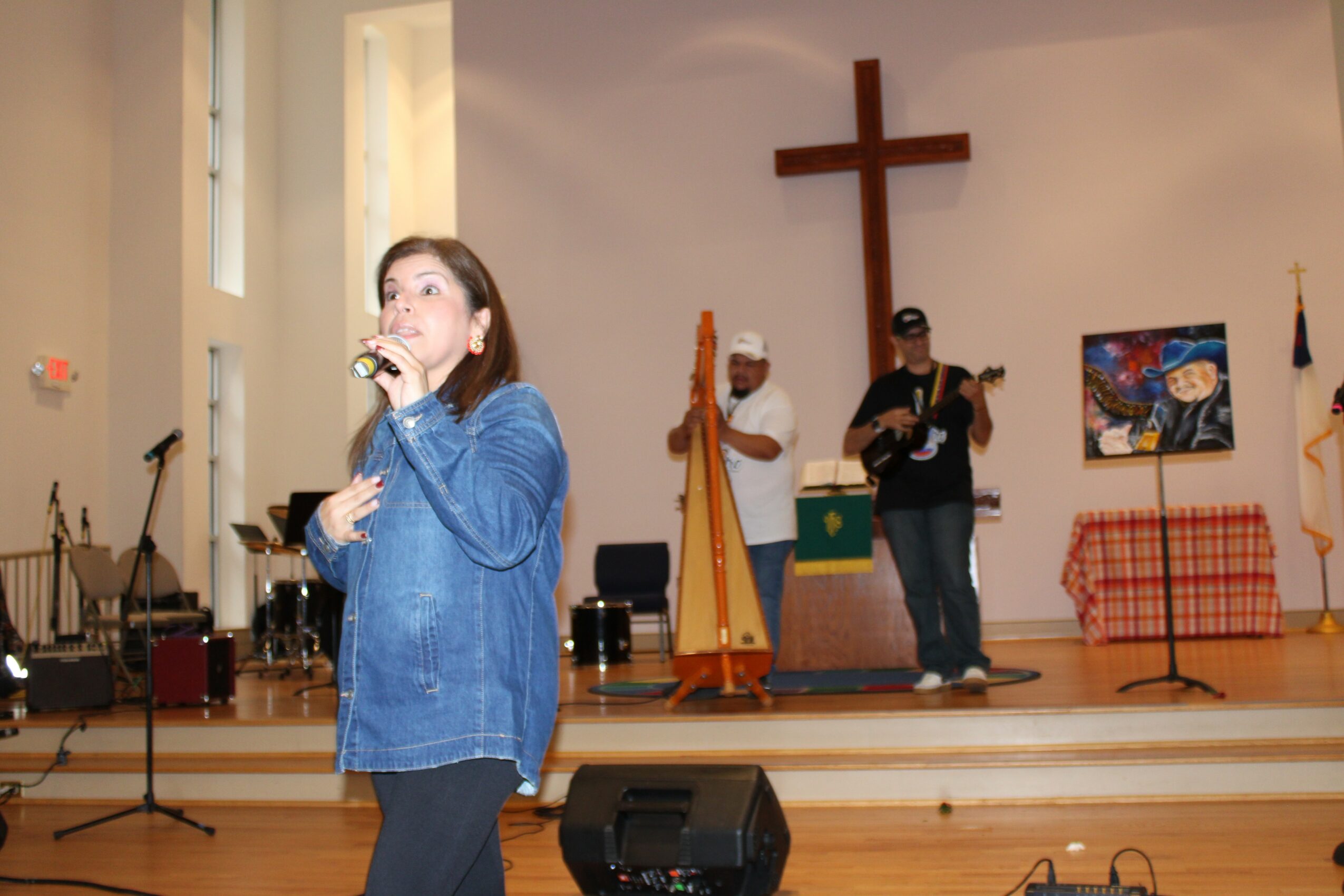 Woman singing on stage with two musicians, a cross, and artwork in the background, in a church setting.