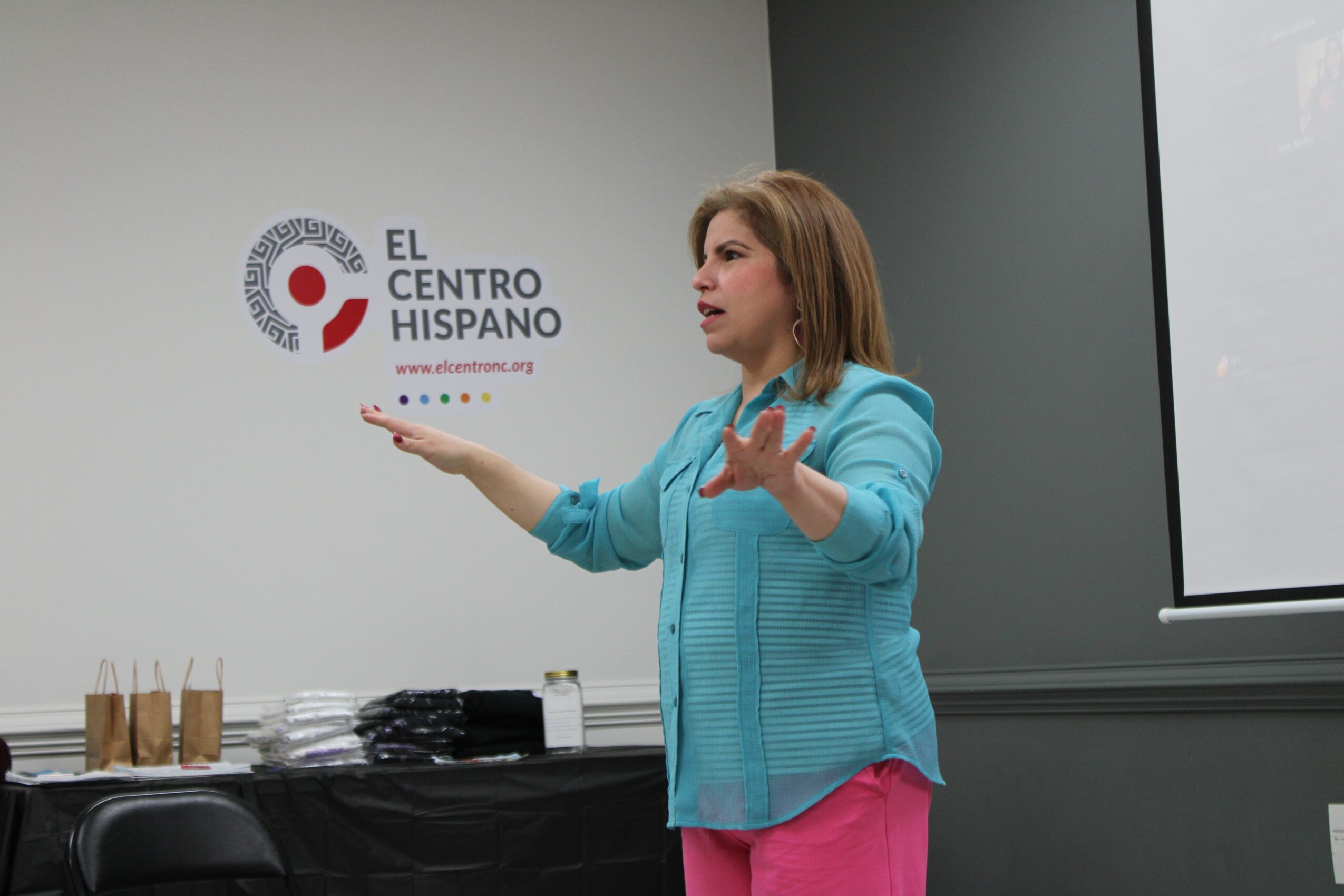 Woman speaking in front of a presentation screen, gesturing with hands, standing near a table with books and papers.