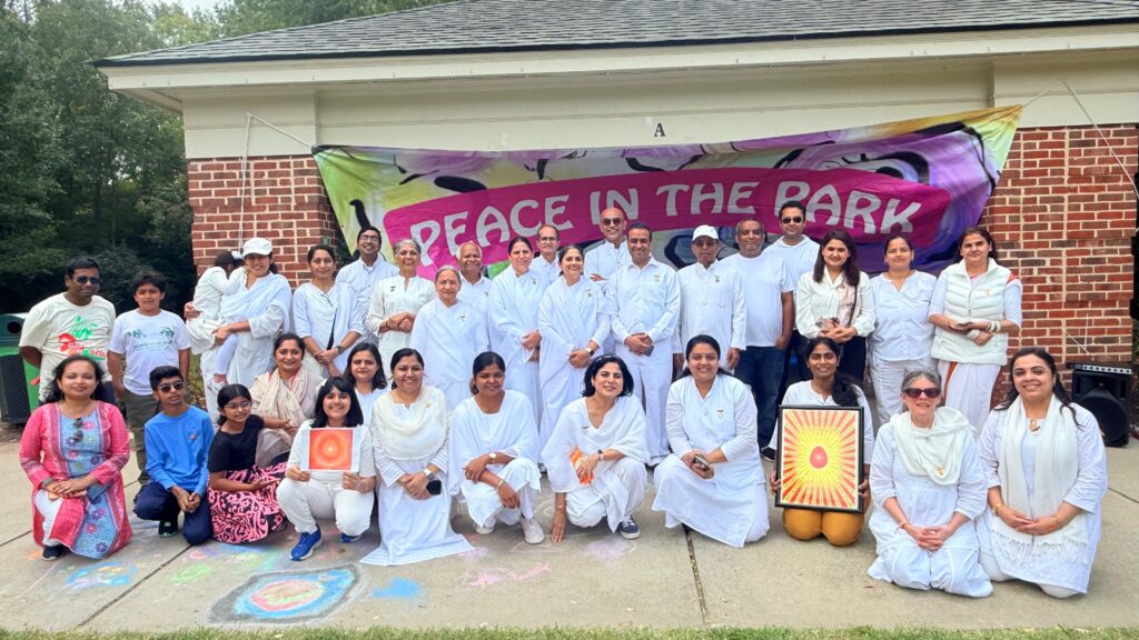 Group of people in white clothing posing outdoors in front of a building with a banner that reads 'Peace in the Park.'