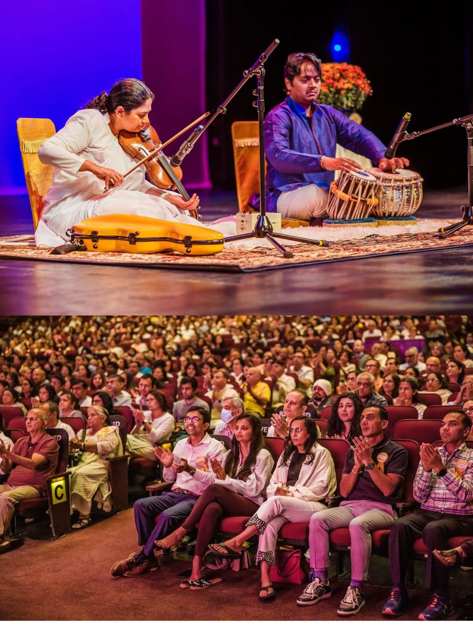 Two musicians perform on stage with instruments, and an audience watches attentively in a large auditorium.