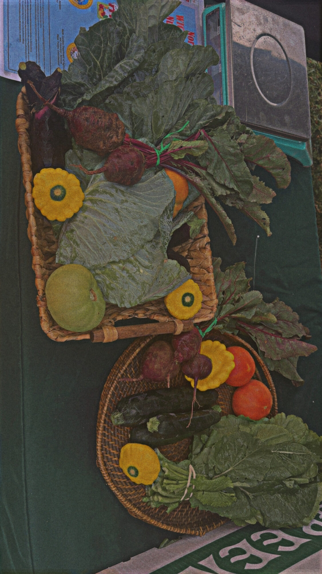 Basket of leafy greens, yellow flowers, and vegetables on a green surface, with a box and electronic device in background.