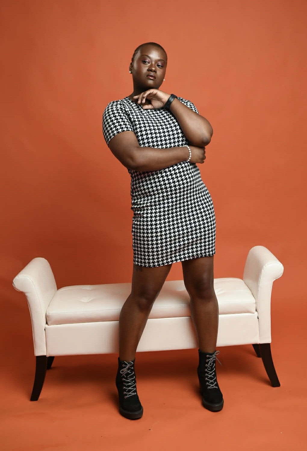 Person standing with arms crossed in front of a white bench against an orange background, wearing a checkered dress and black boots.