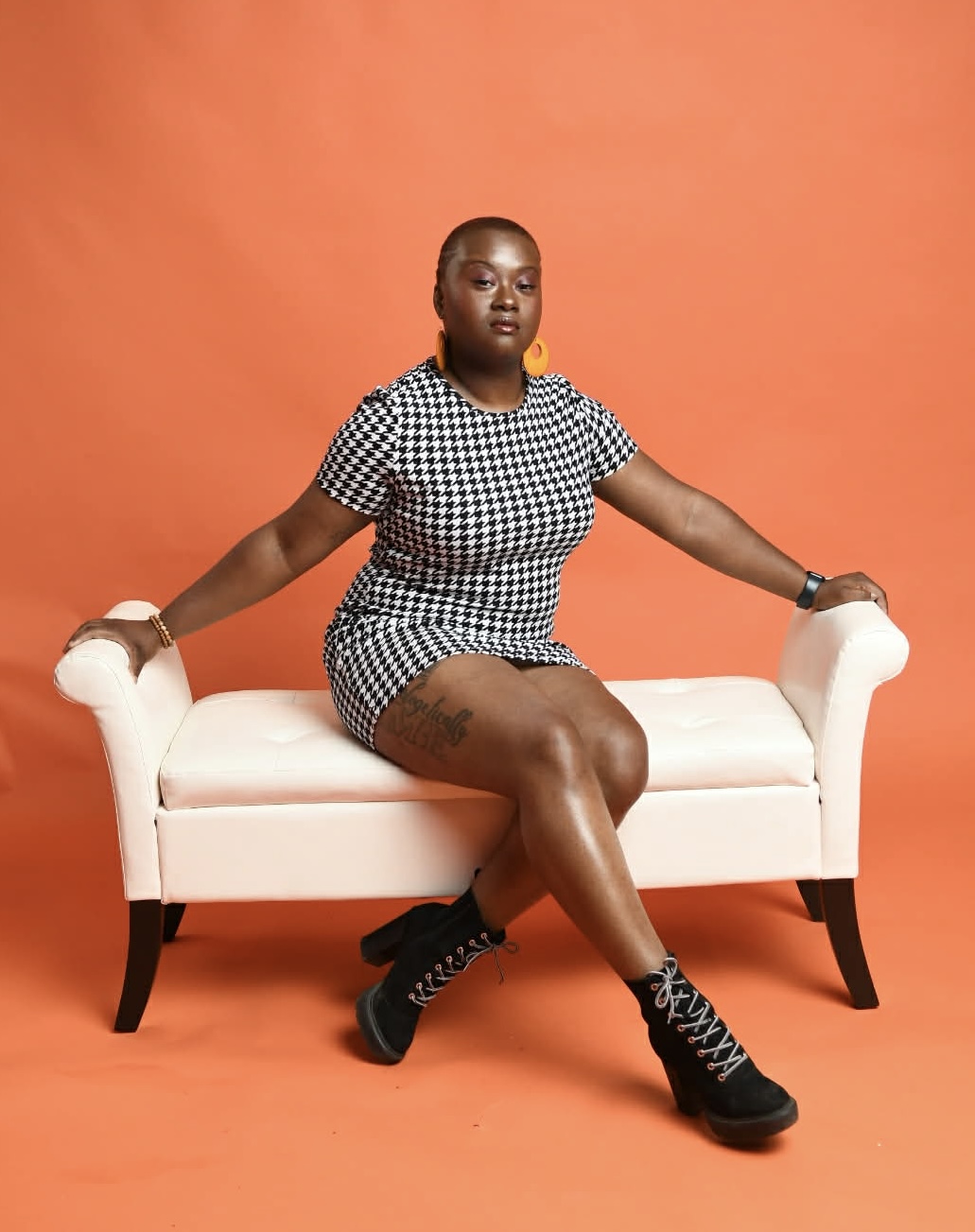 Person sitting on a white bench against an orange background, wearing a checkered outfit and black boots.