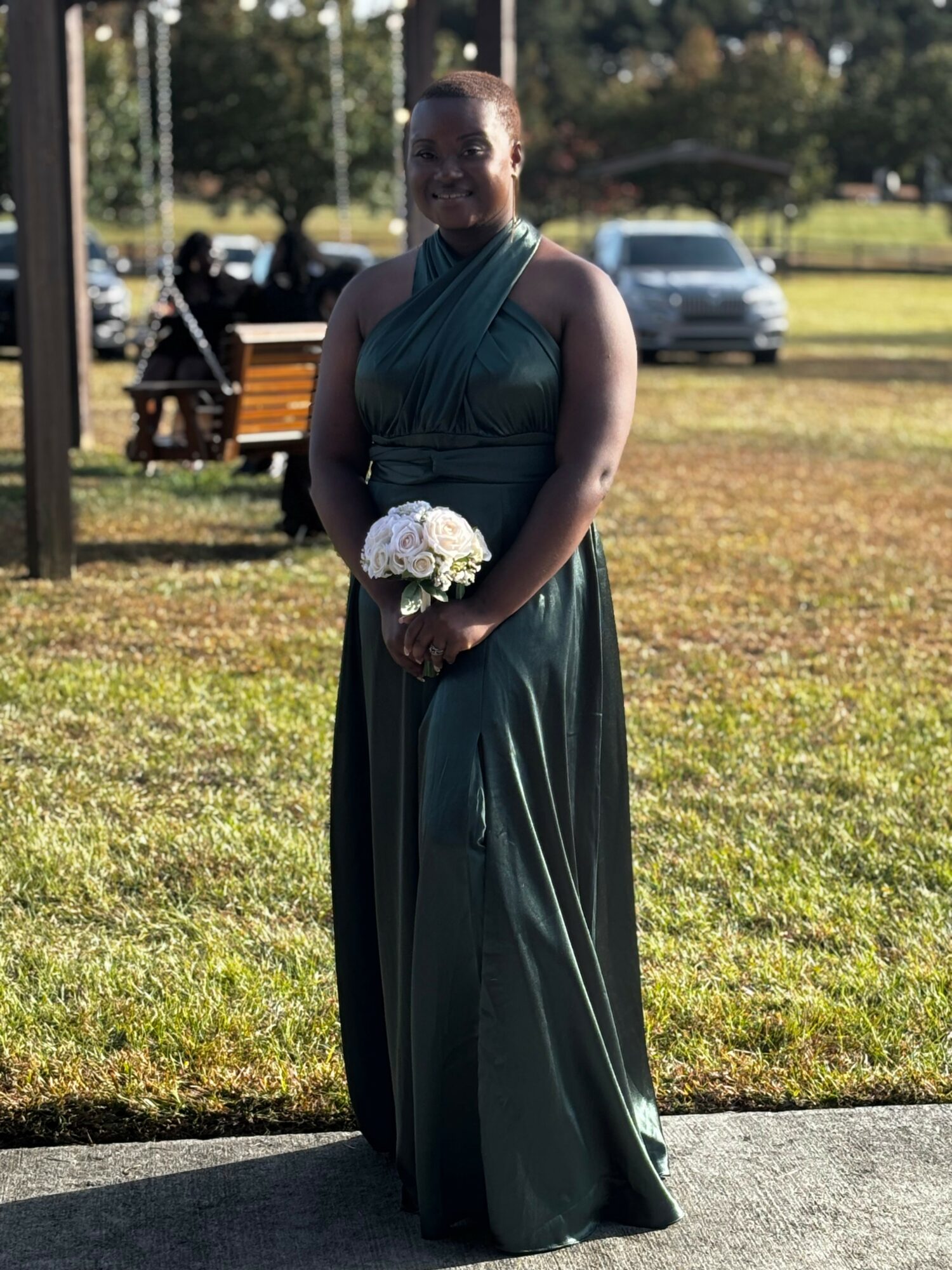 Woman in a dark green dress holding a bouquet of white flowers outdoors, with trees and parked cars in background.