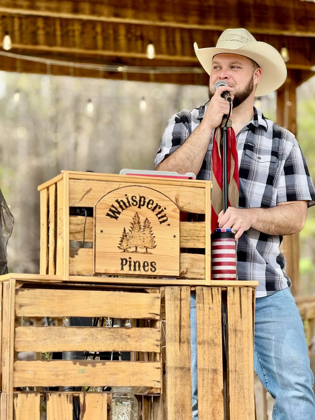 Man wearing a cowboy hat and plaid shirt speaking into a microphone outdoors, with wooden crates and a thatched roof in background.