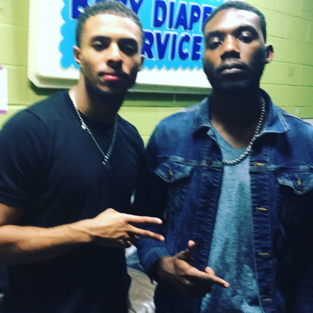 Two young men standing indoors, one making a peace sign, the other with a serious expression, in front of a sign.