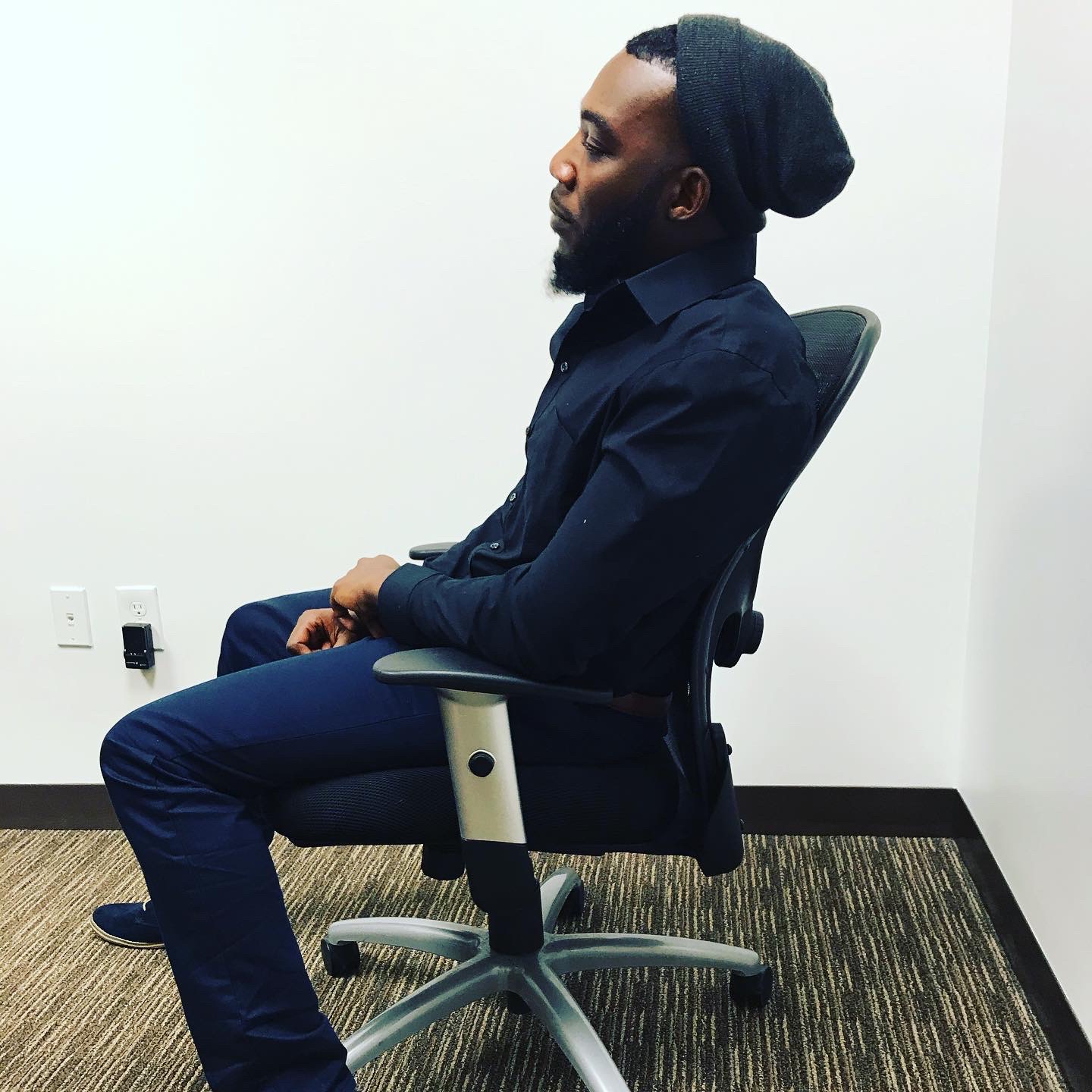 Side view of a man sitting in an office chair against a white wall, wearing a dark shirt and beanie.