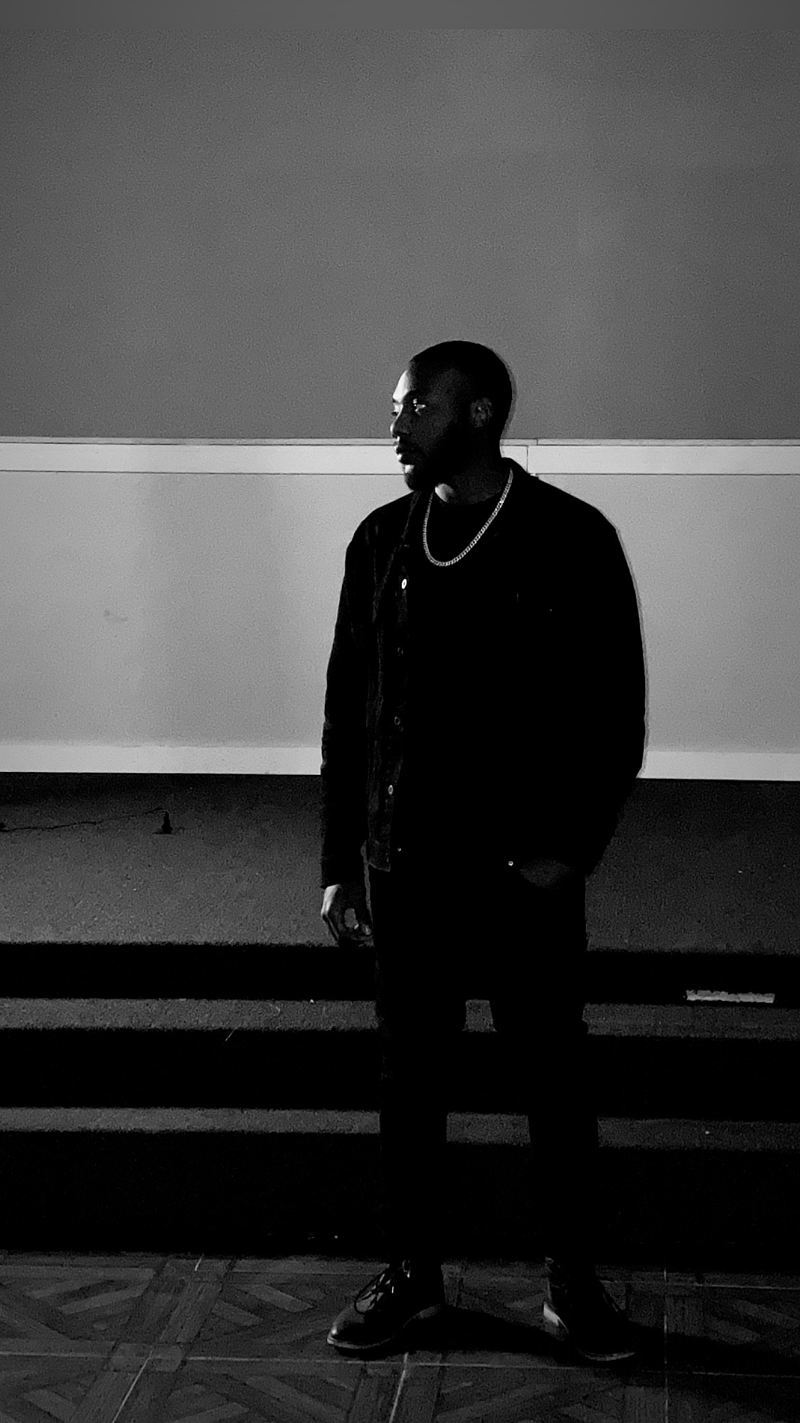 Person standing in front of a wall, facing left, wearing dark clothing and glasses, in a dimly lit environment.