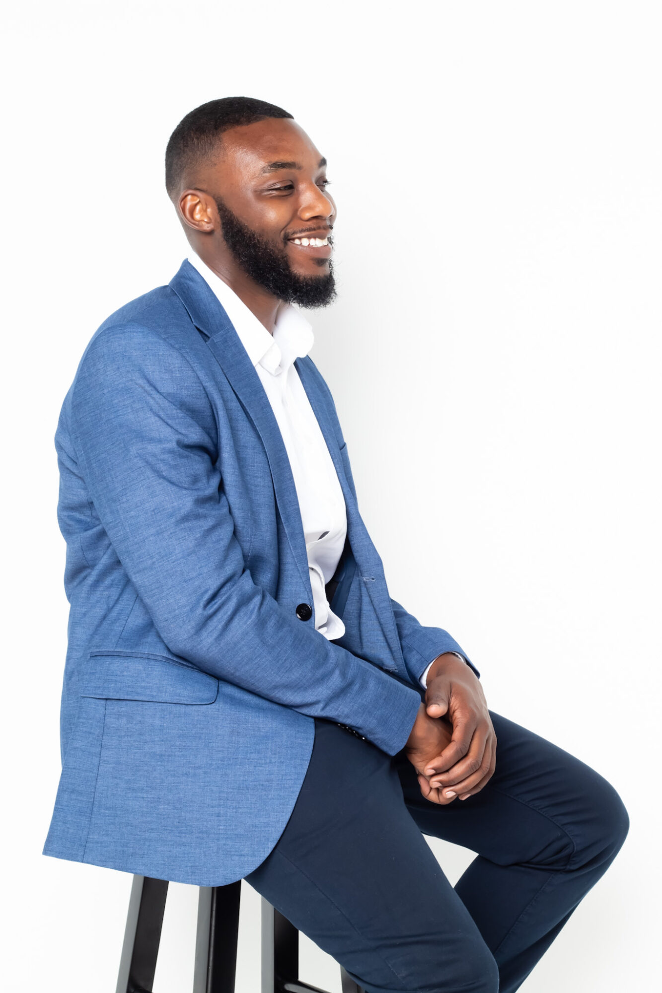Man in blue blazer and white shirt sitting on a stool, smiling, against a white background.