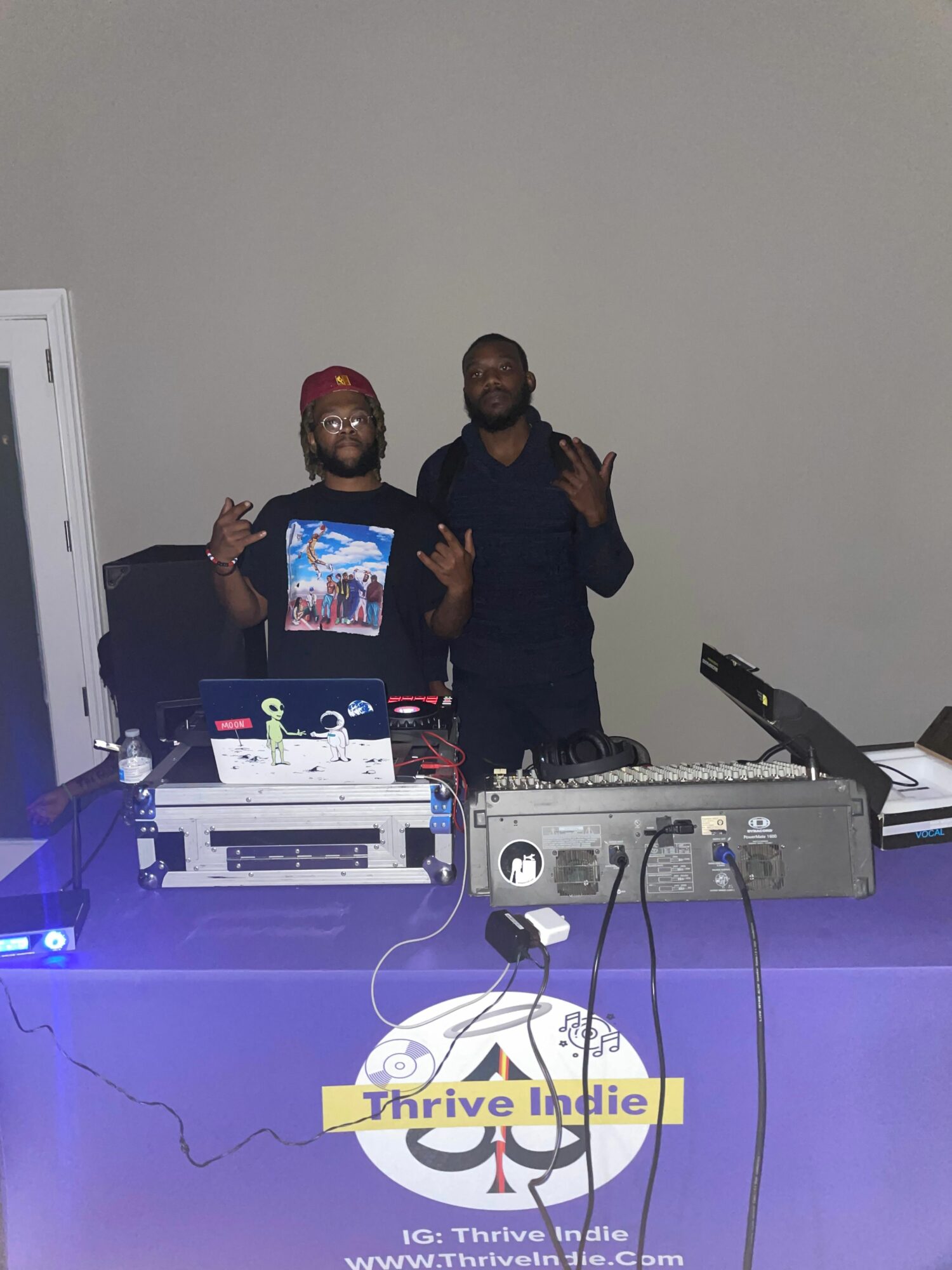 Two people standing behind DJ equipment, one making a peace sign, in a room with a plain wall.