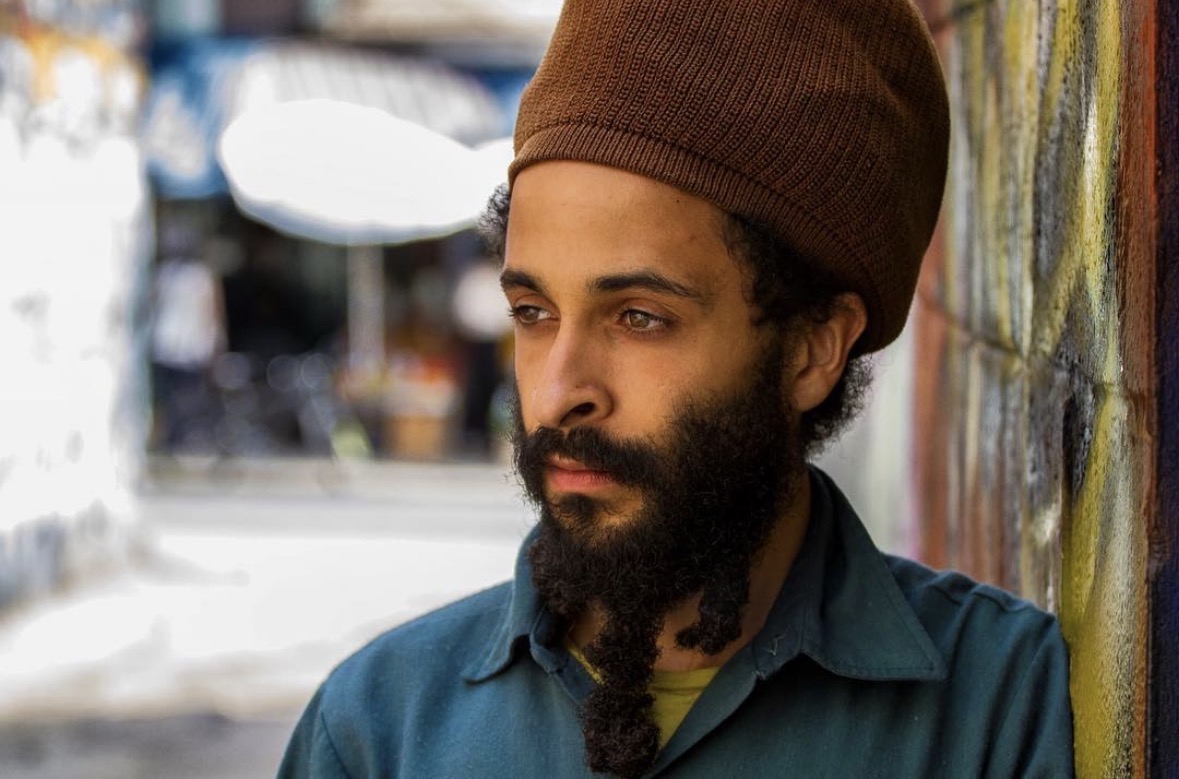 Man with a beard and brown beanie leaning against a colorful wall outdoors.