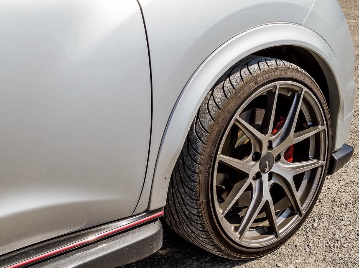 Close-up of a car's front wheel with a black alloy rim and red brake caliper, part of a silver vehicle.