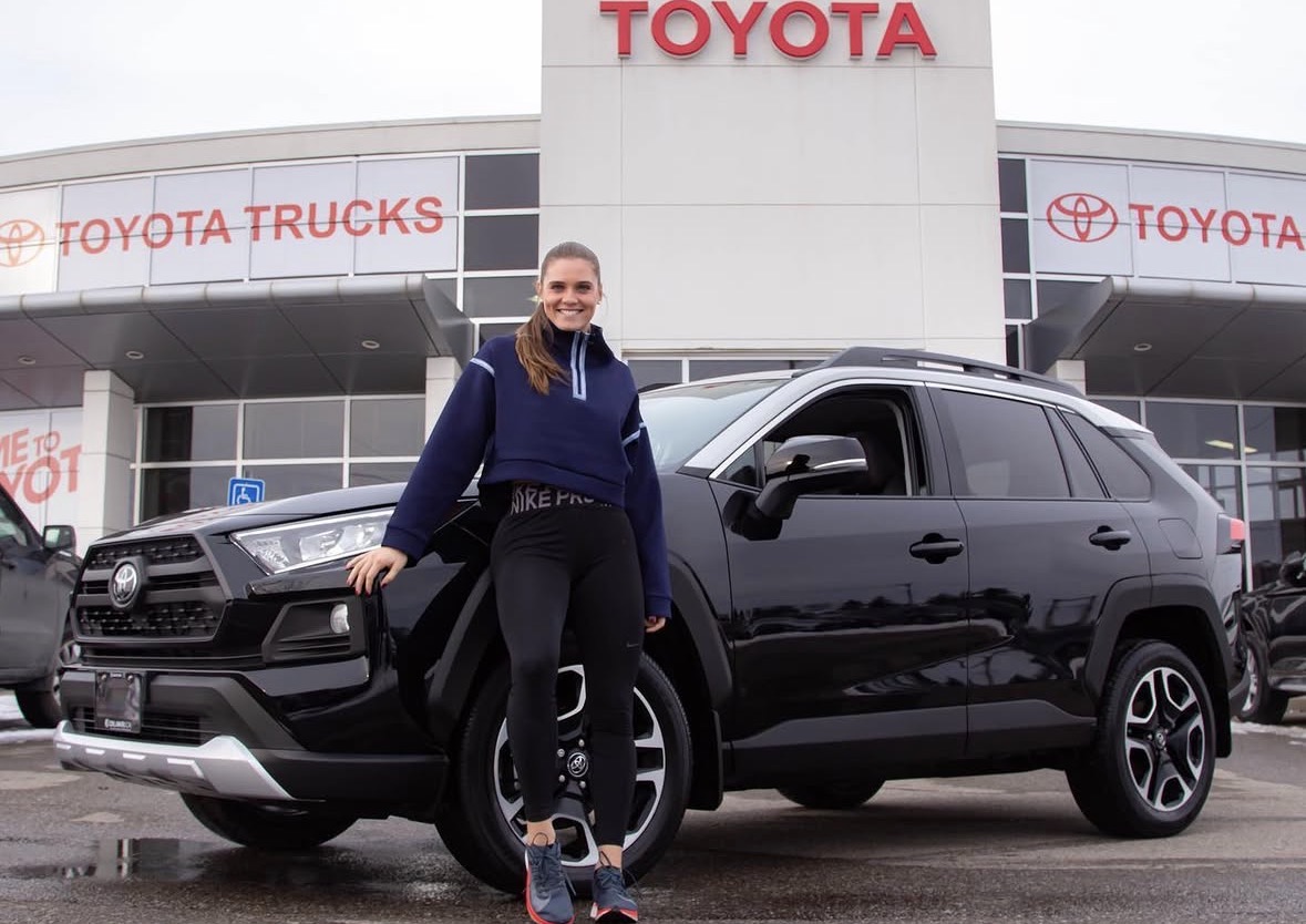 Person standing next to a black SUV in front of a Toyota dealership building with Toyota signs.