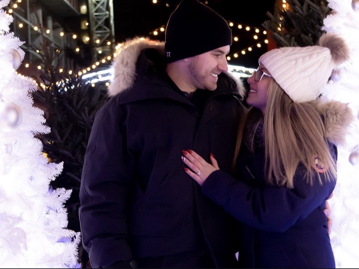 A couple in winter clothing smiling and looking at each other outdoors at night with Christmas lights and decorated trees.