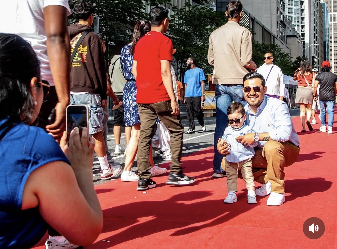 Man and child pose for photo on red carpet with city buildings in background.