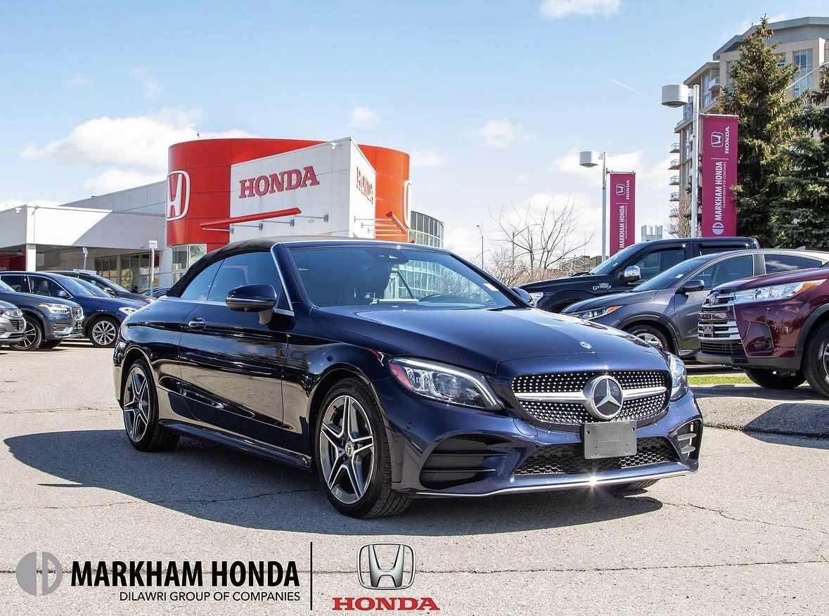 Black Mercedes-Benz car parked in a dealership lot with Honda and other cars, dealership signs, and buildings in background.