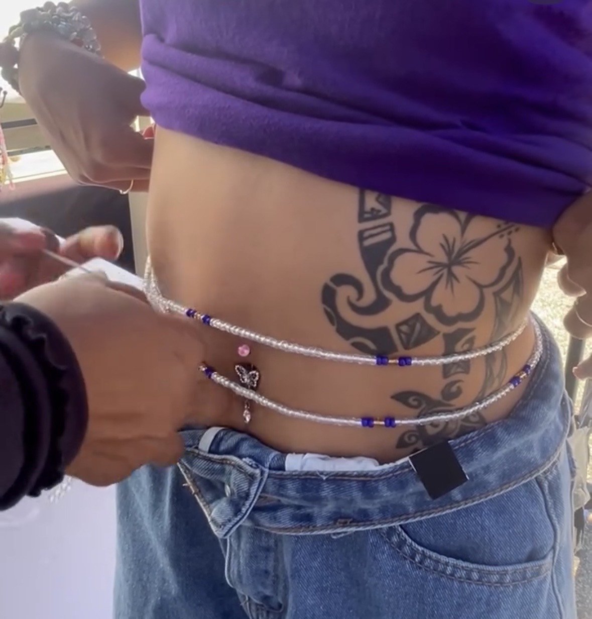 Close-up of a person's midsection showing a tattoo, a purple shirt lifted, and a beaded waist chain.