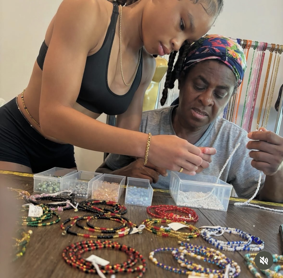 Two women examine jewelry pieces on a table, with colorful beads and jewelry displayed in front of them.