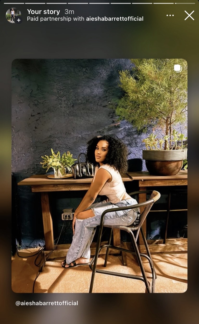 Woman with curly hair sitting at a wooden table in a room with a dark textured wall and plants, looking at the camera.