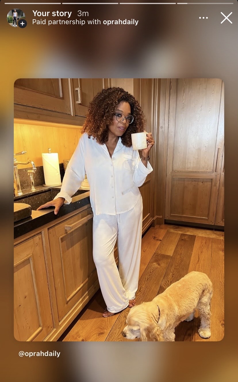 Woman with curly hair in pajamas holding a mug, standing in kitchen with a dog nearby, wooden cabinets and floor.