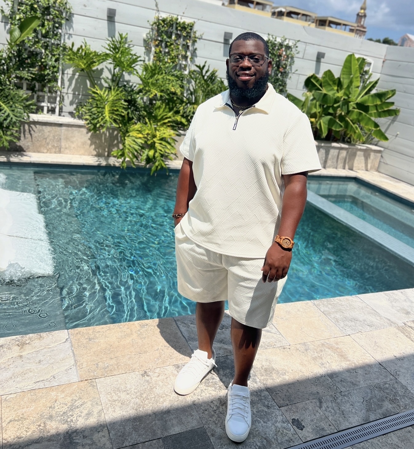 Man standing by a swimming pool outdoors, wearing a white outfit and sneakers, with plants and buildings in background.