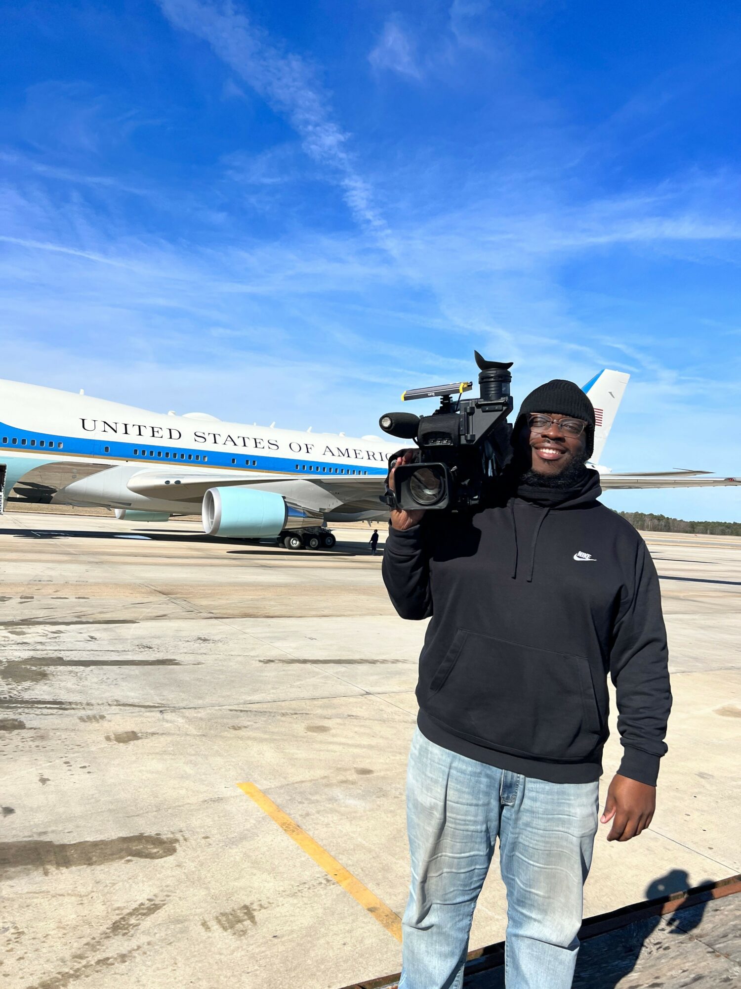 Person holding a camera on a tarmac with a plane in the background, under a blue sky.