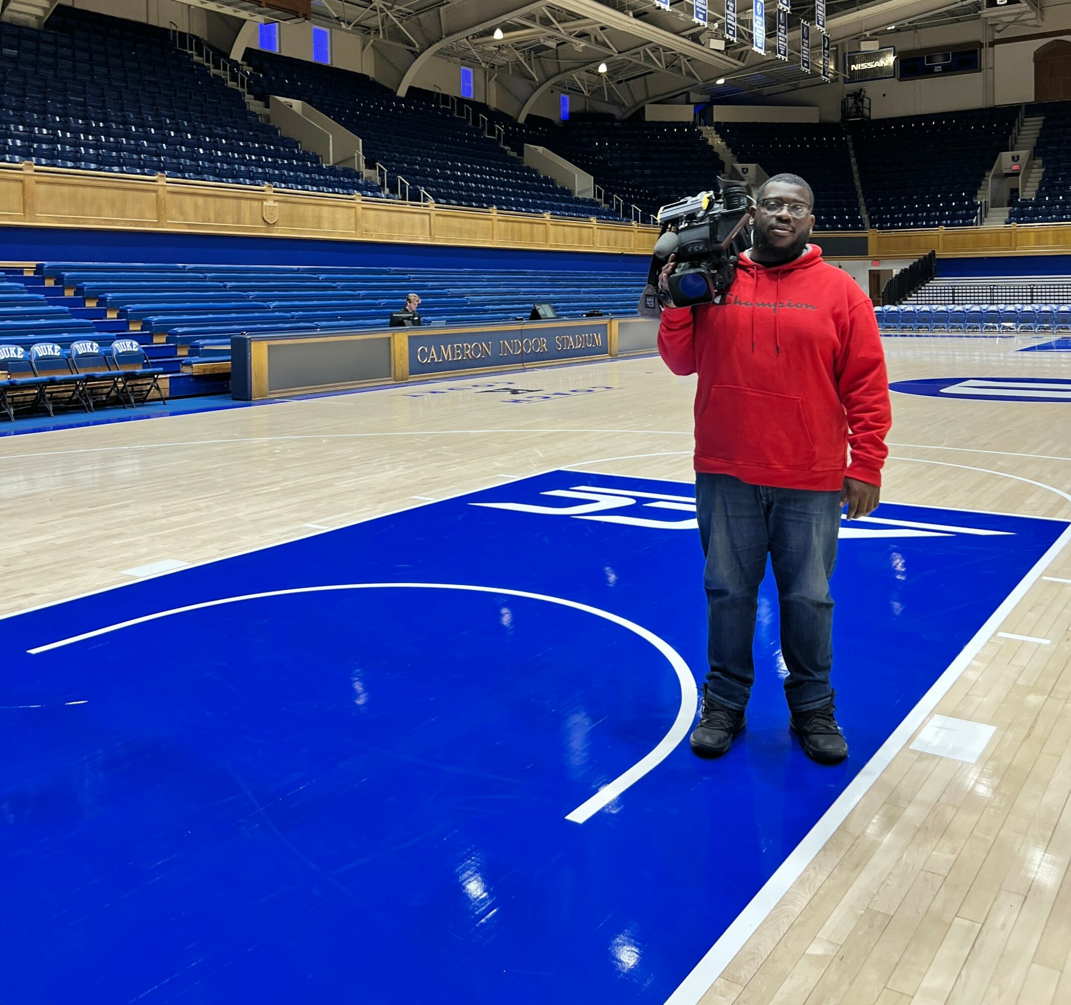 Person in red hoodie holding a camera on a basketball court with blue and white markings, empty seats in background.