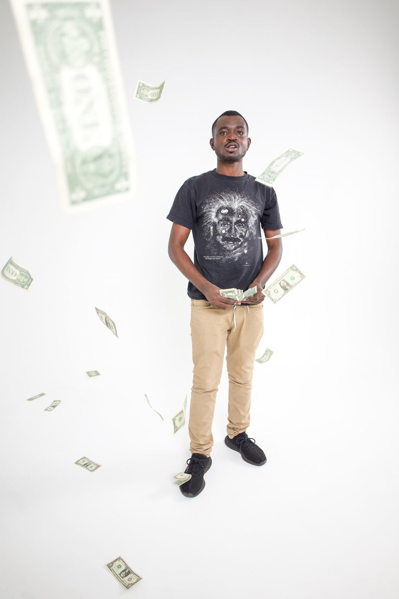 Young man standing with hands in pockets, surrounded by falling dollar bills, against a plain white background.
