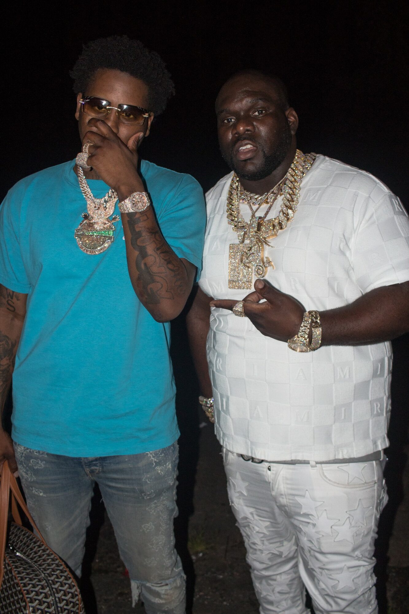 Two men stand together, wearing jewelry and casual clothing, against a dark background.