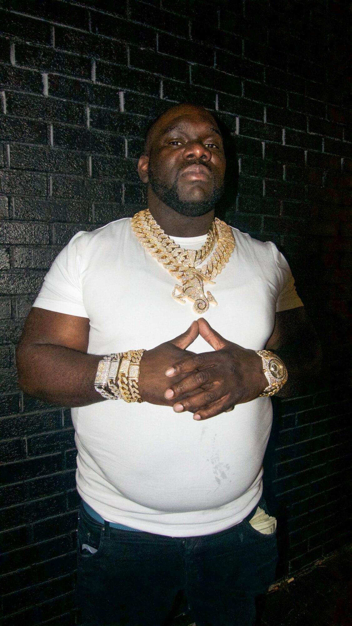 Man with gold jewelry and white t-shirt standing against a dark brick wall, hands clasped in front.