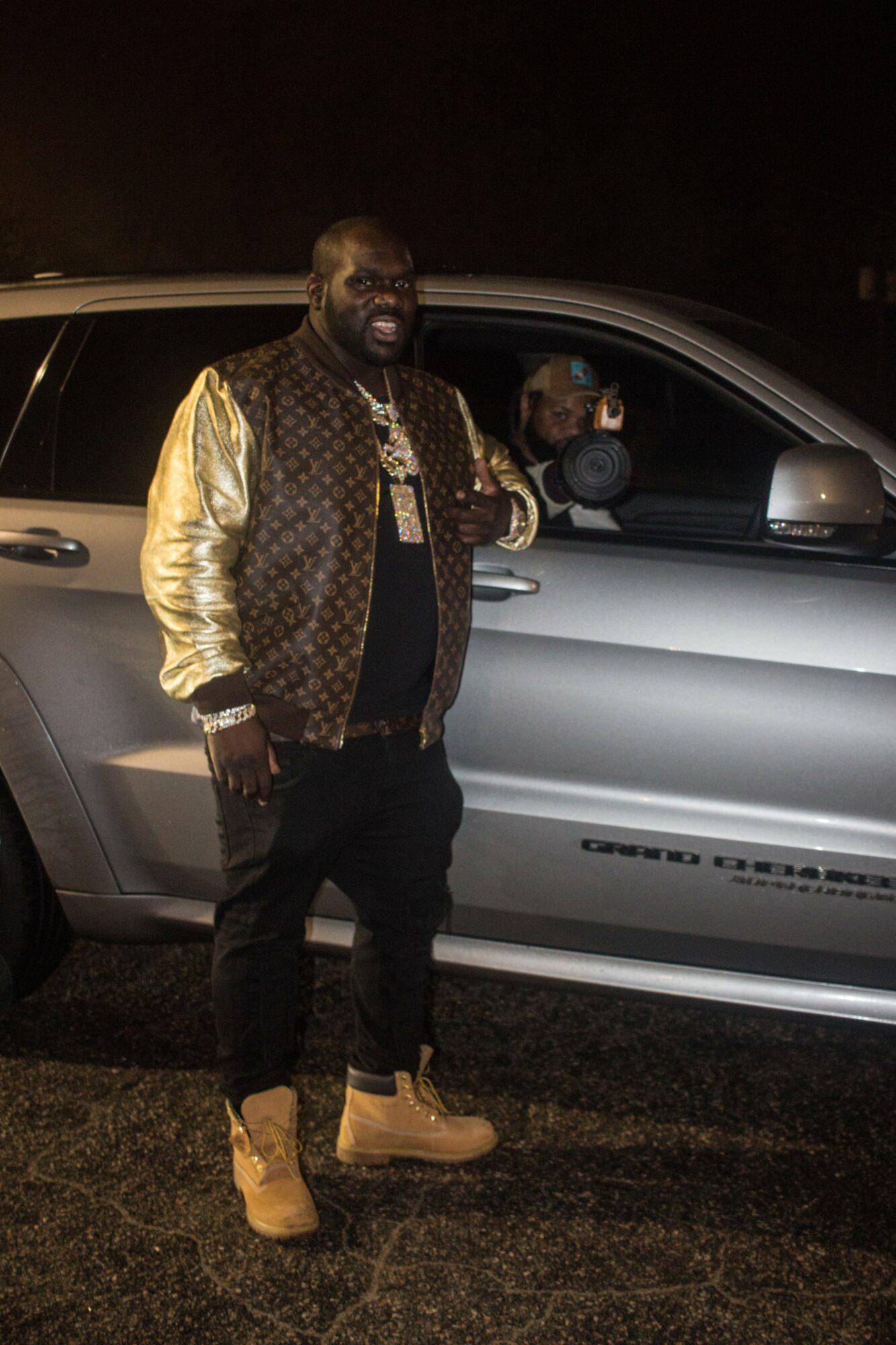 Man standing next to a silver car at night, smiling, wearing a gold and black jacket, black pants, and tan boots.