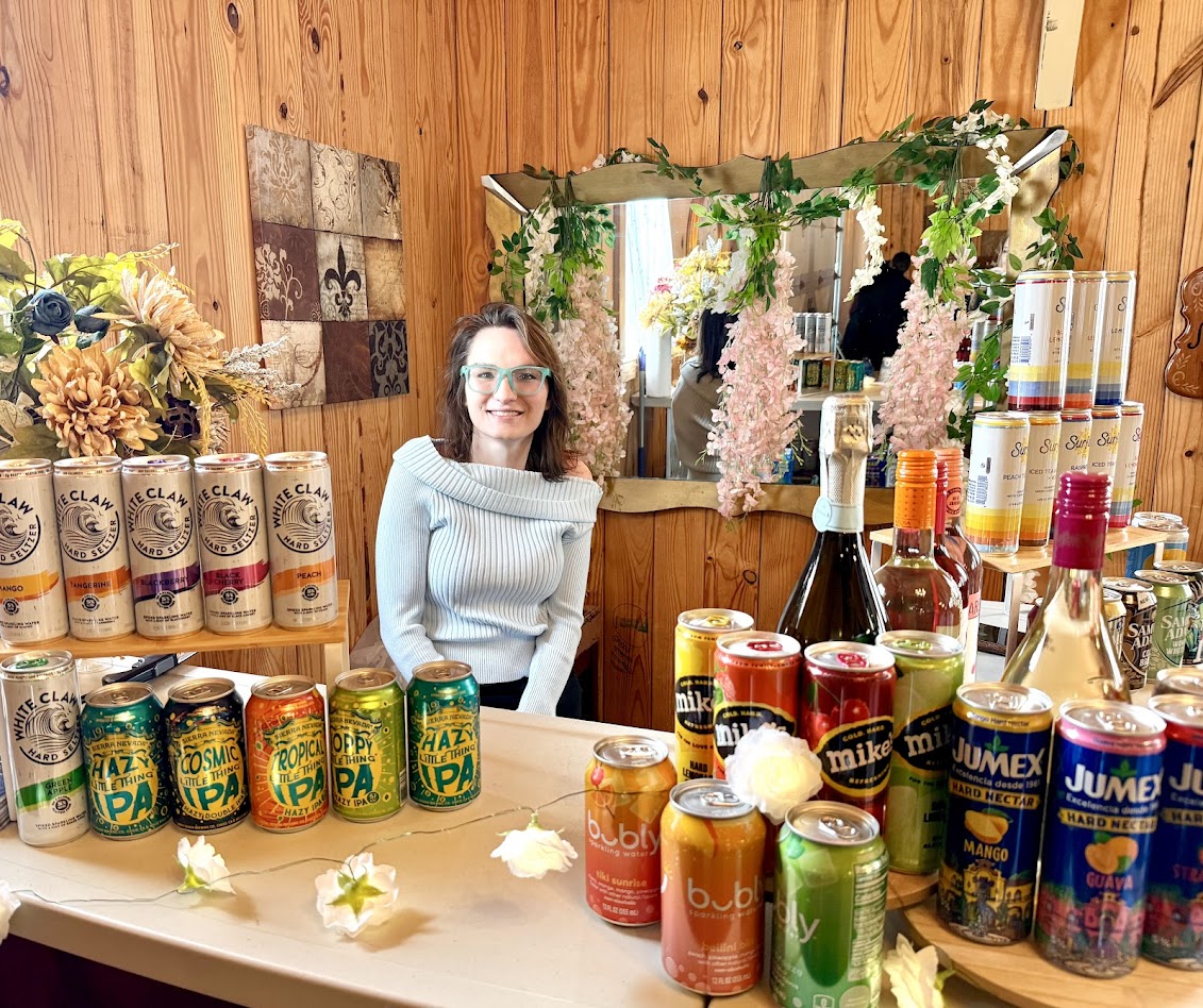 Woman with glasses and curly hair behind a table with drinks, flowers, and decorations in a wooden room.