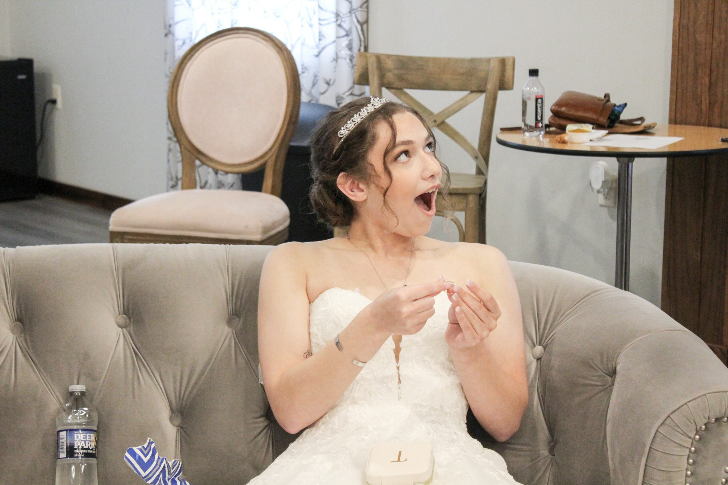 Woman in wedding dress sitting on sofa with surprised expression, hands near face, in a room with chairs and table
