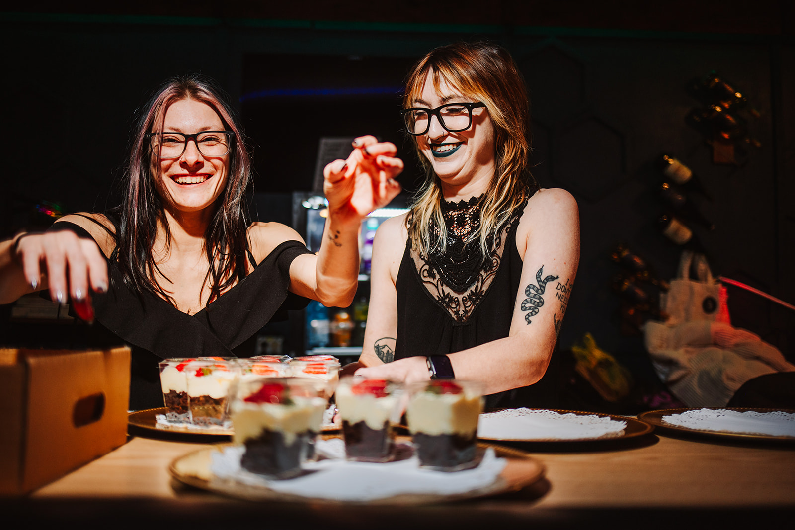 Two women smiling and decorating cakes at a table with various desserts, in a dimly lit room.