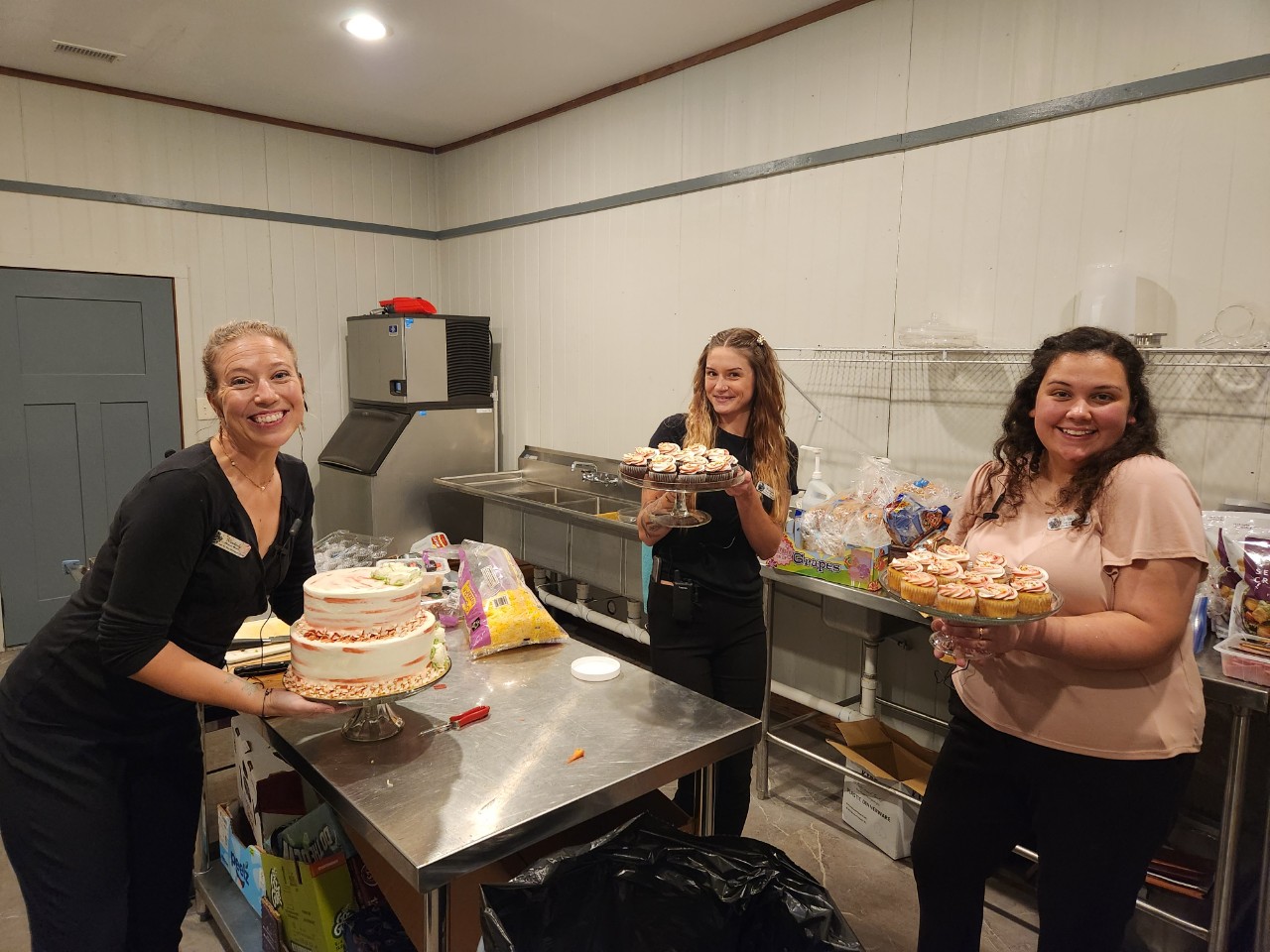 Three women in a room with a table, holding cakes and smiling, with shelves and a door in the background.