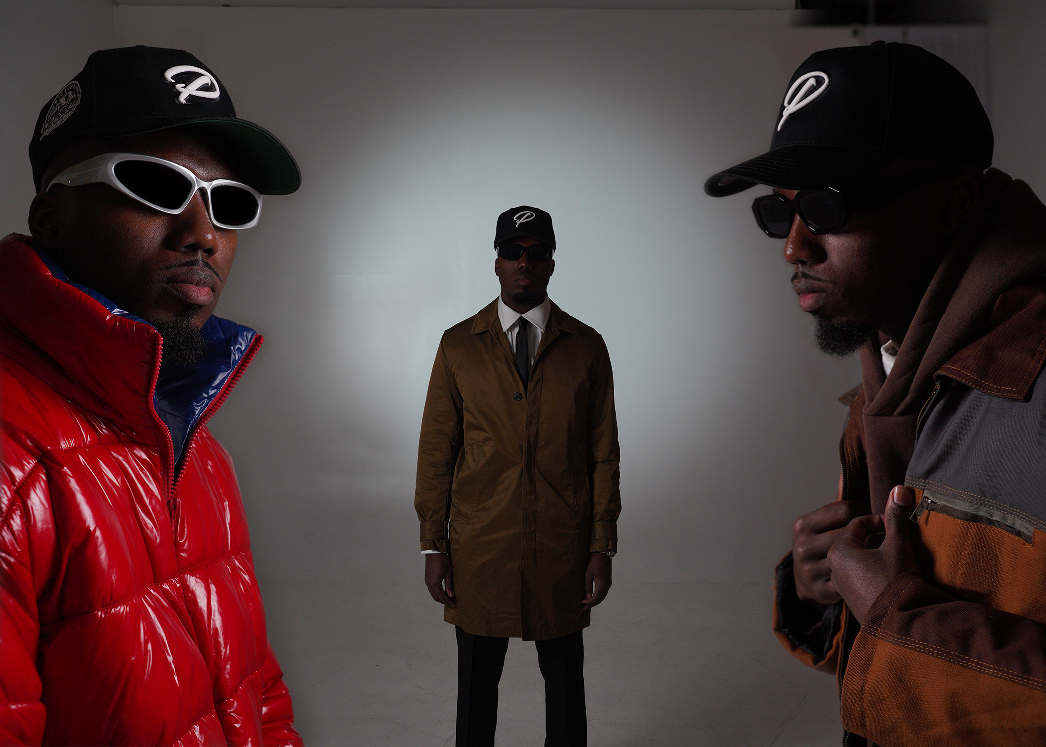 Three men in stylish clothing and sunglasses pose indoors against a plain background.