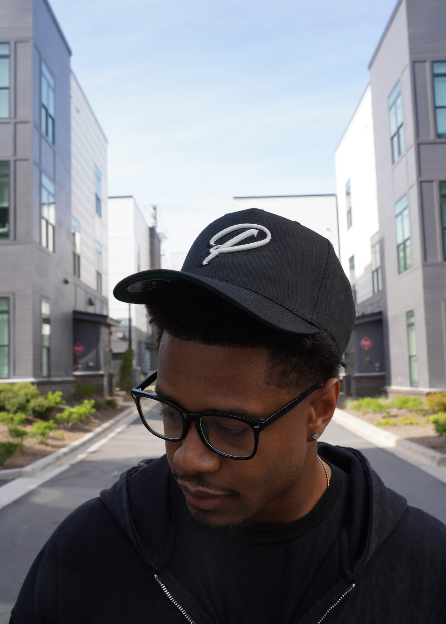 Young man wearing glasses and a black cap outdoors in an urban area with modern buildings.