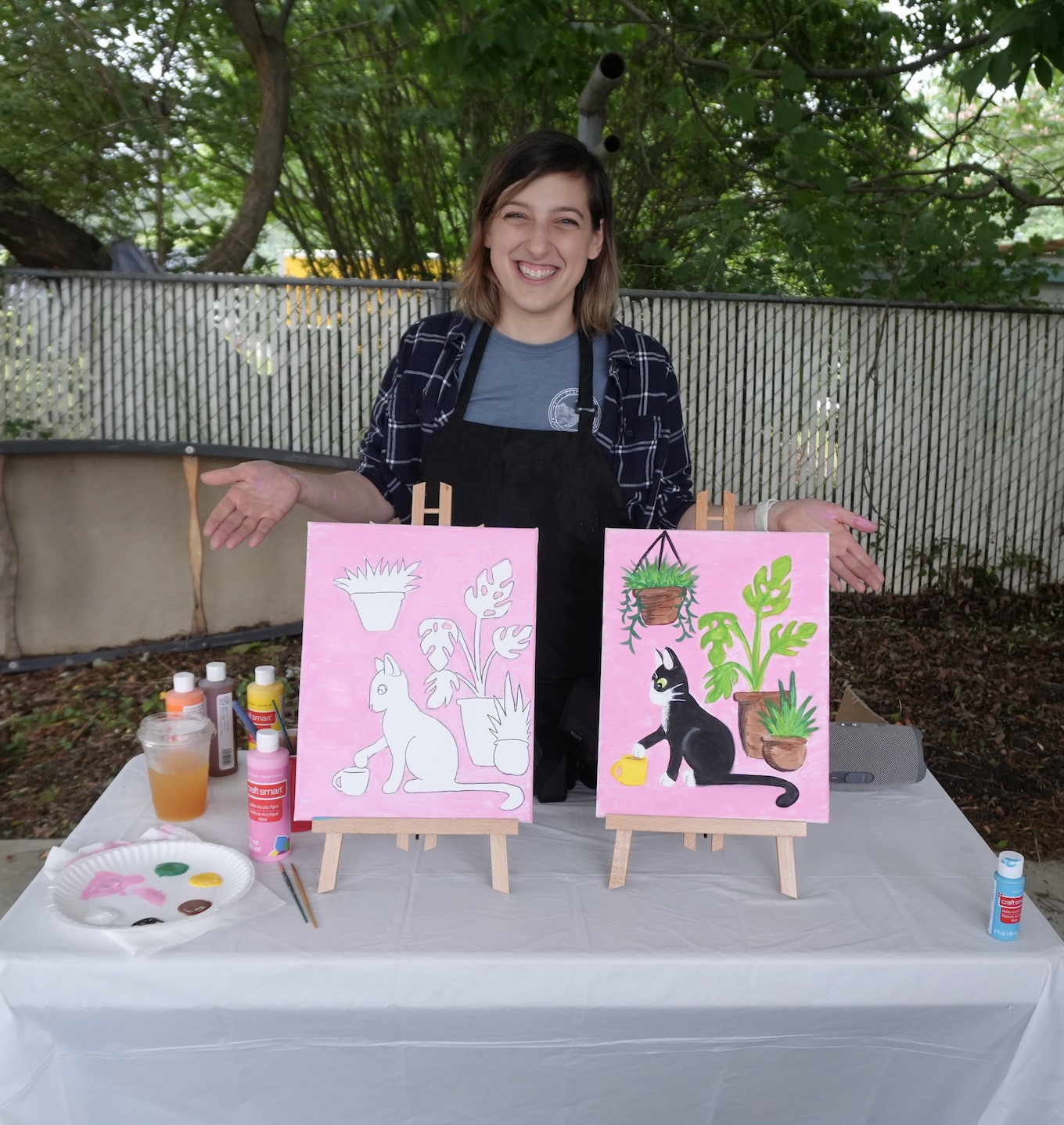 Young woman standing behind a table with two pink paintings of animals, outdoors with trees and a fence in background.