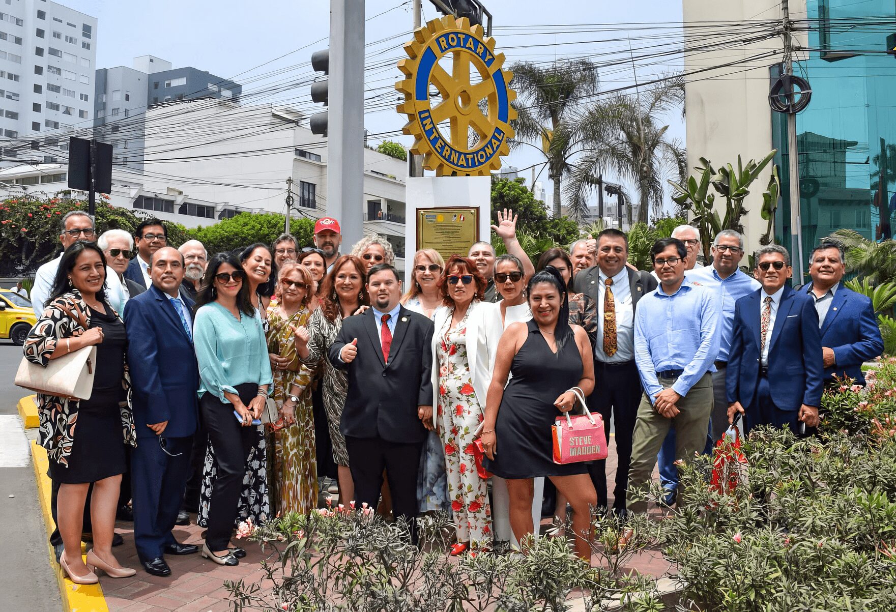 Group of people standing outdoors in front of Rotary International sign, some waving, with buildings and palm trees in background.